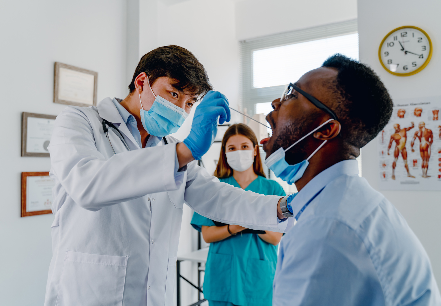 Black man having his throat swabbed by a male doctor of Asian descent for a PCR test at medical clinic as a White female nurse looks on.