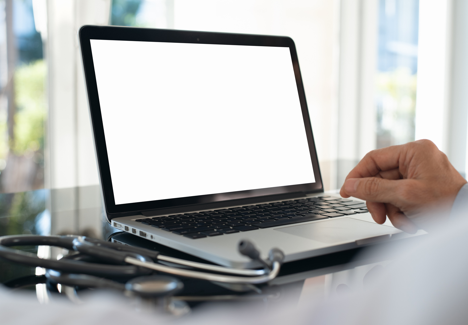 Doctor working on blank screen laptop computer in modern doctor office with stethoscope and notebook on desk. iStock image.