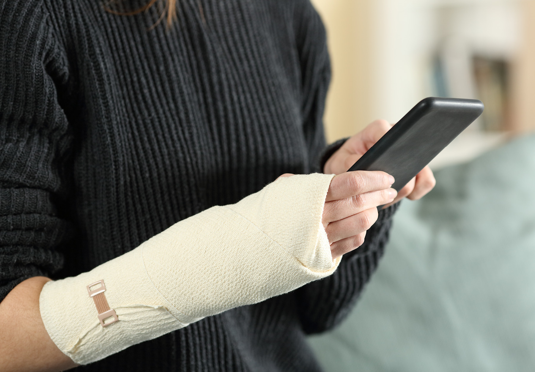 Close up of an injured woman's hands with bandaged arm using mobile phone at home. She is wearing a black sweater. iStock image.
