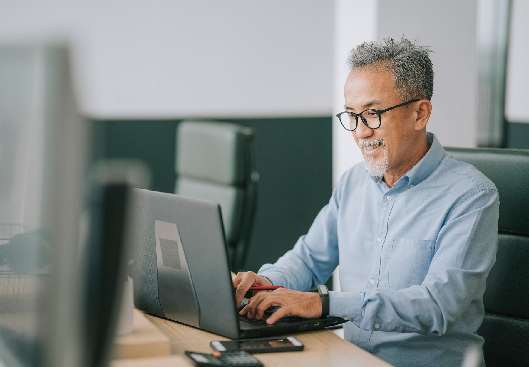 A middle-aged man of Asian descent types on a computer in an office. He is wearing glasses and smiling. iStock image.