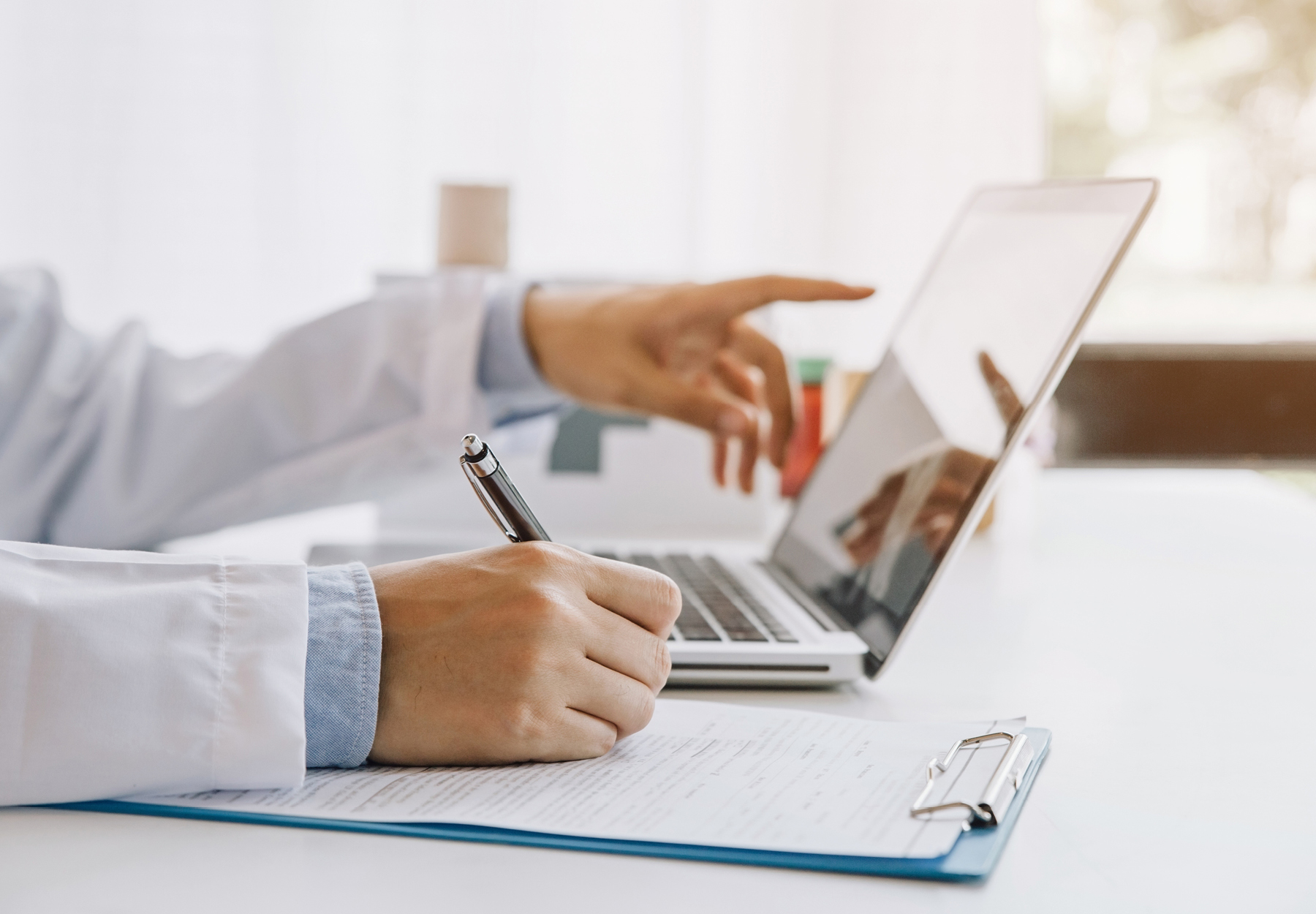 Closeup of a lab professional's hands working at a desk with a laptop, while writing something on a clipboard. iStock image.