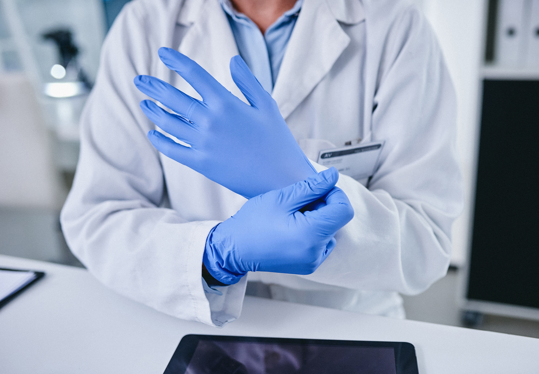 Closeup of lab professional putting on gloves while standing above a desk with a tablet. iStock image.