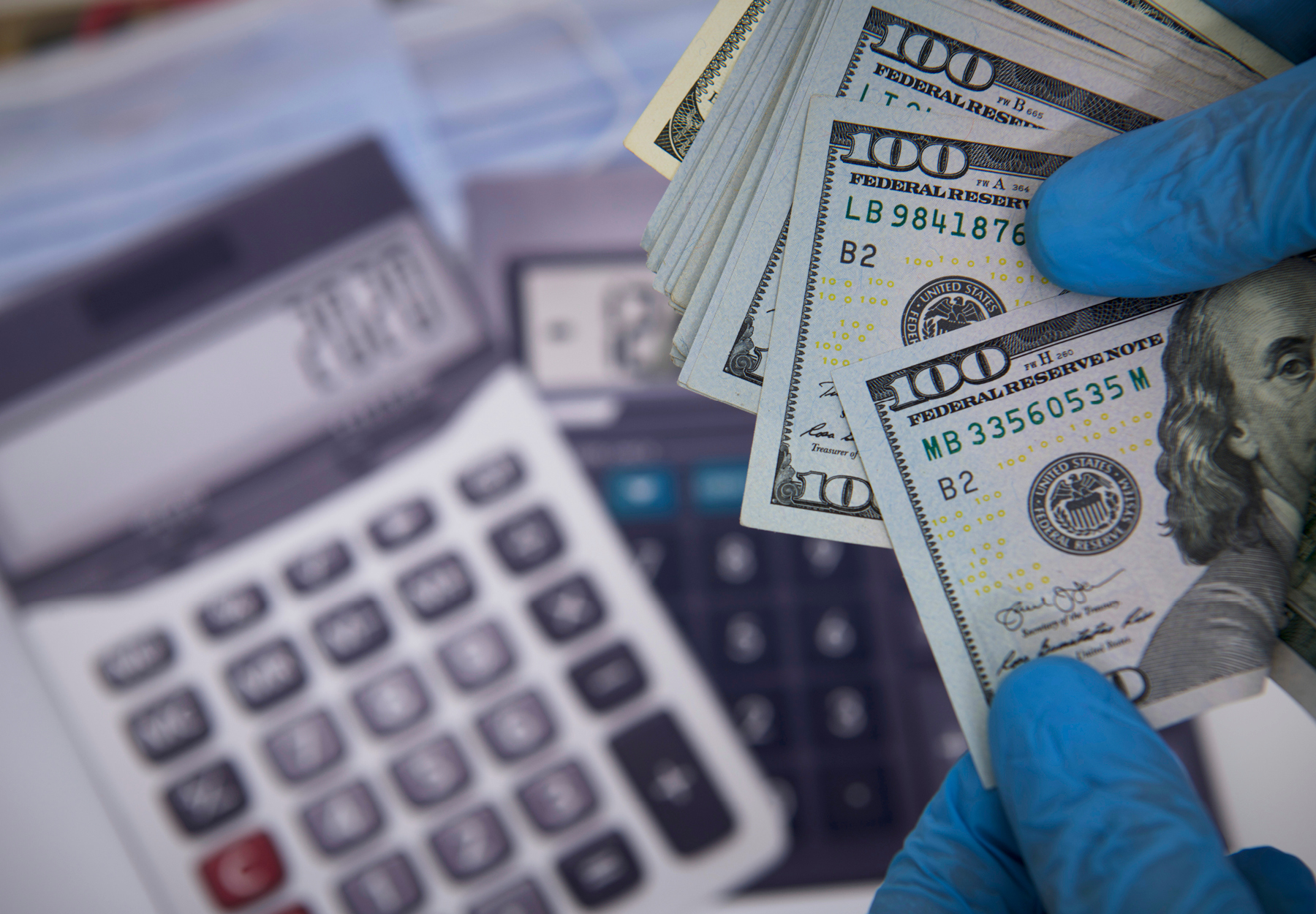Closeup of blue gloved hand of lab professional holding US bills over calculators. iStock image.
