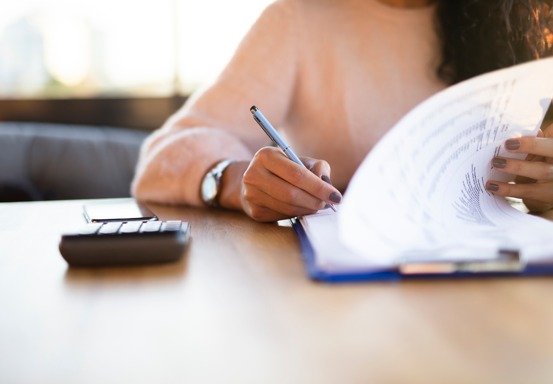 Closeup of woman in long-sleeved pink shirt filling out health insurance form on a blue clipboard. iStock image.