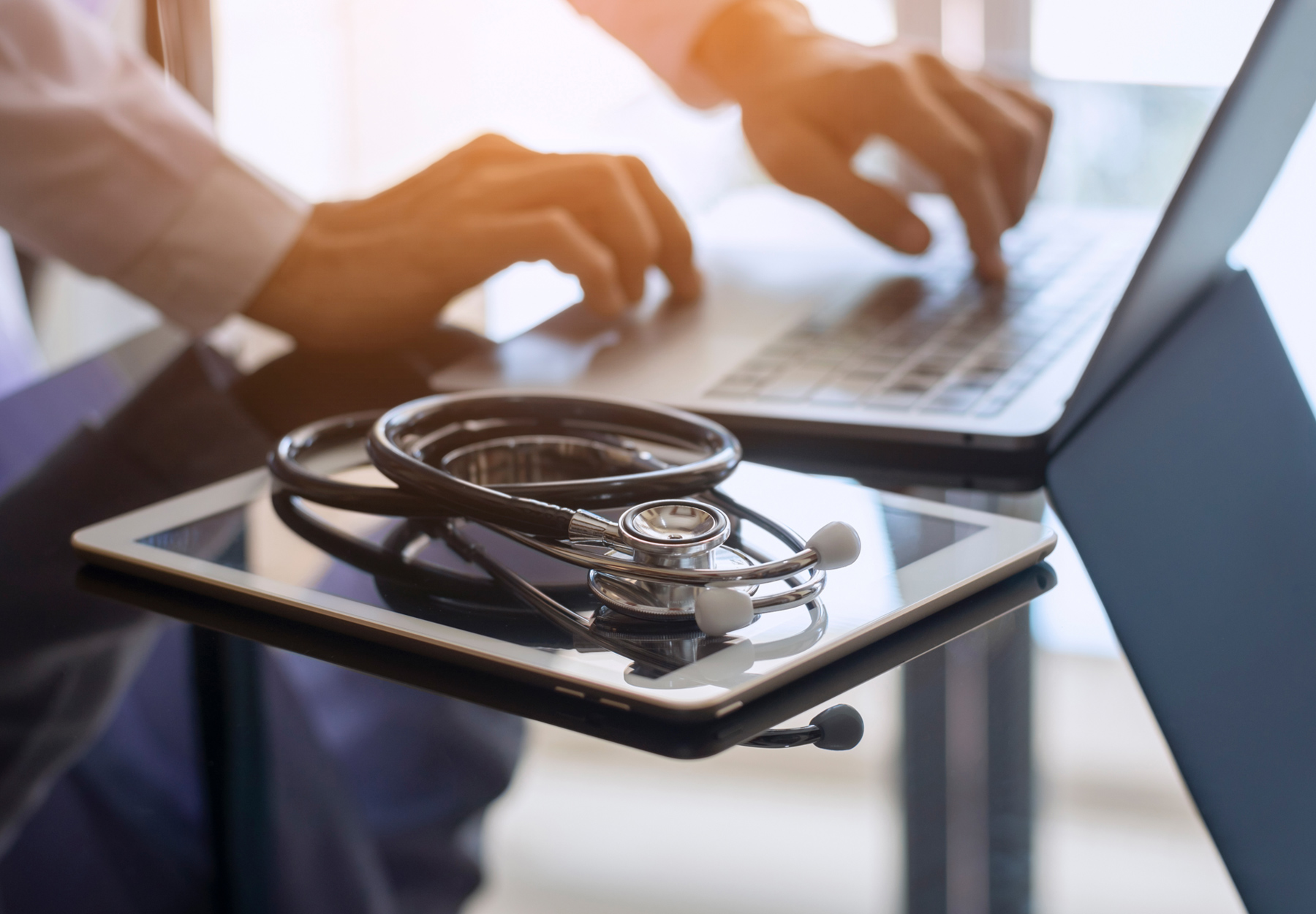 Closeup of doctor using a laptop with a tablet and stethoscope on the desk nearby. iStock image.
