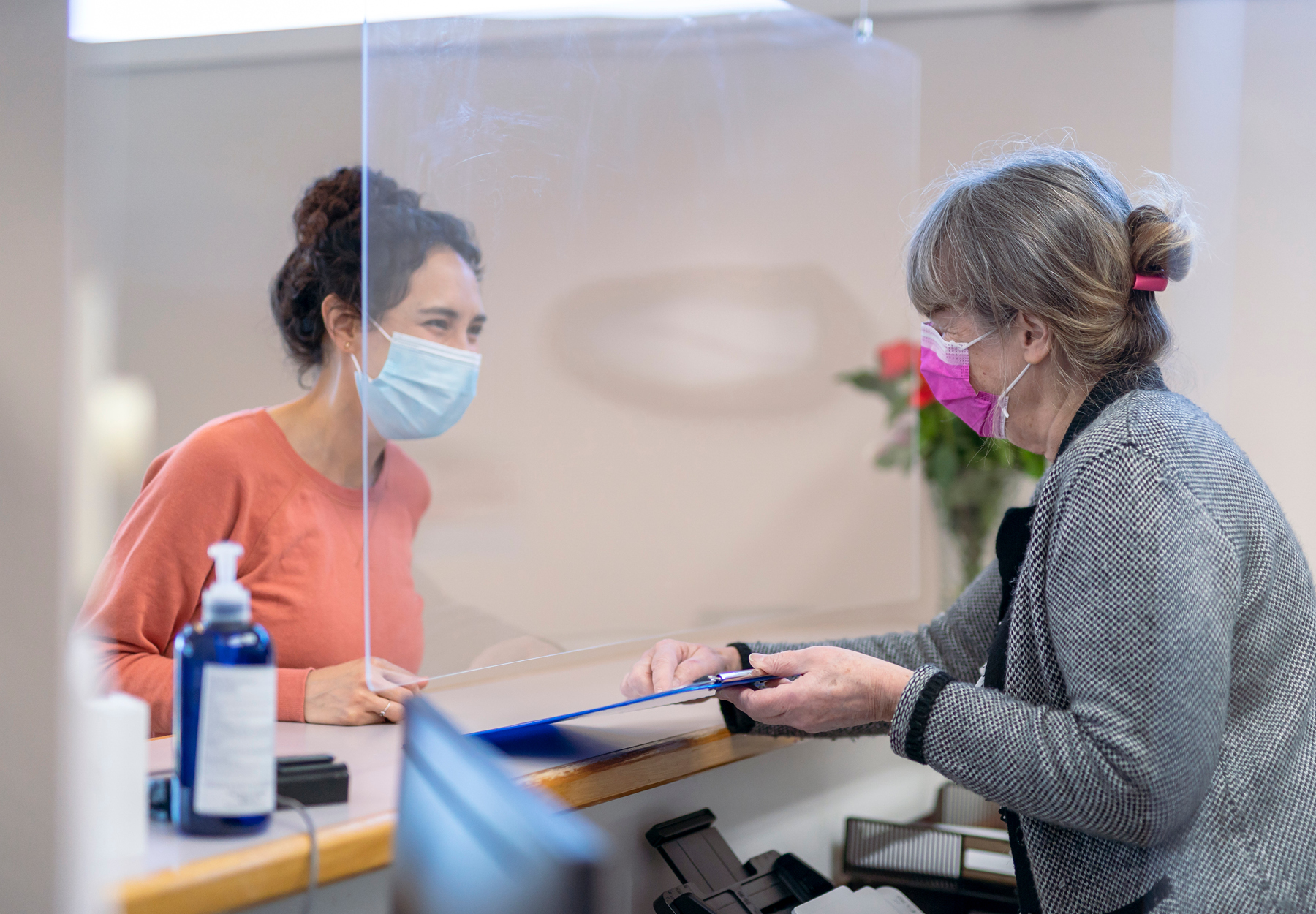 Masked female receptionist at a healthcare clinic gives a clipboard with a form to a masked female patient. Stock image.