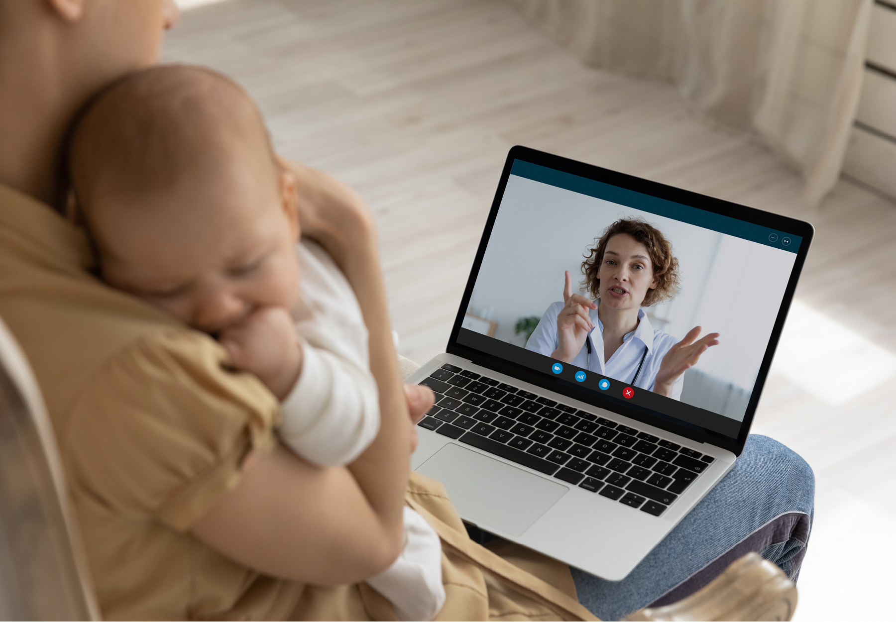 Mother holding baby while doing a telehealth call on her laptop with her doctor. iStock image.