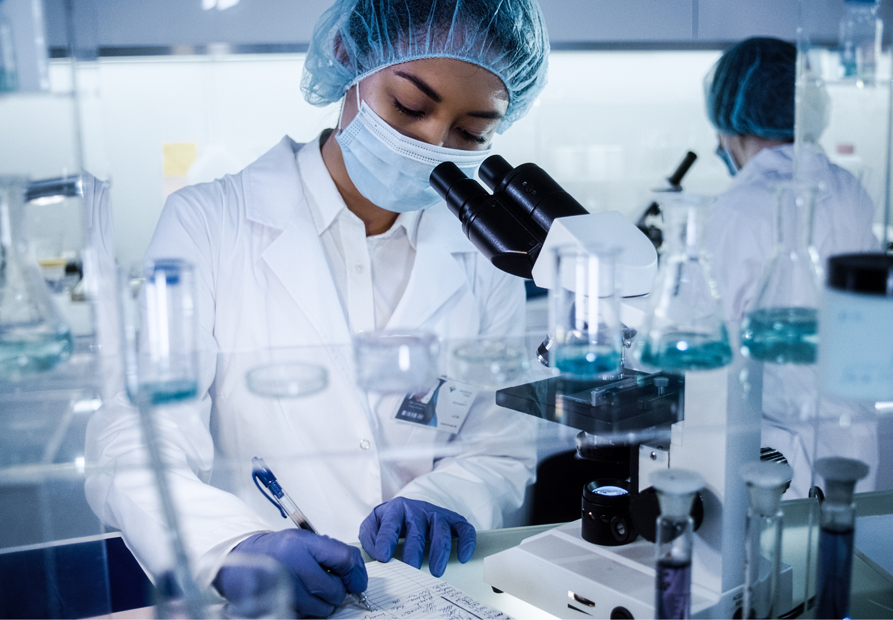 Female lab employee in full PPE stands in front of a microscope and makes notes in a notebook. iStock image.