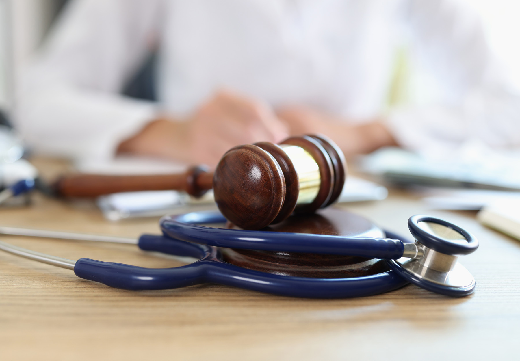 Closeup of judge's gavel and stethoscope with doctor in background writing notes. Concept of legal aspects of healthcare. iStock image.