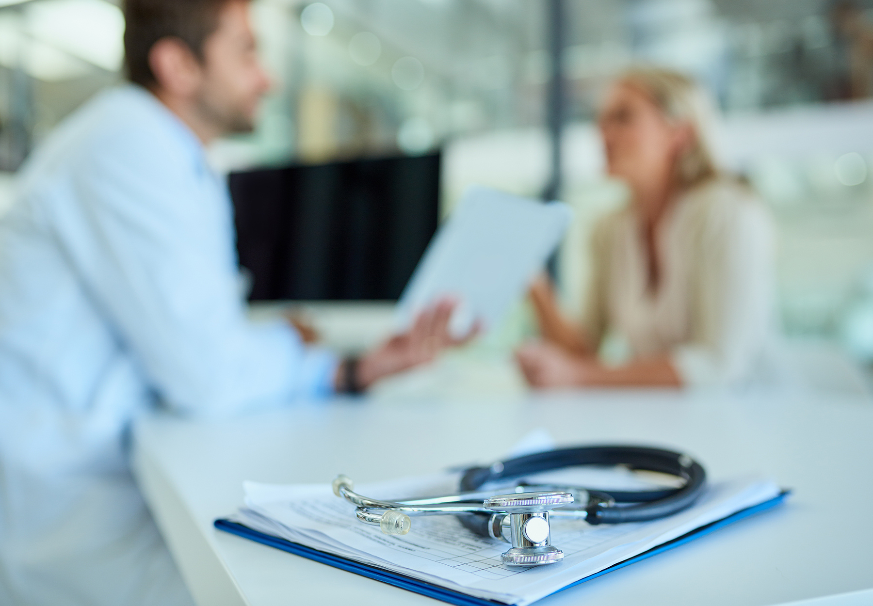 Shot of a stethoscope and a clipboard on a desk with a doctor and patient in the background. iStock image.