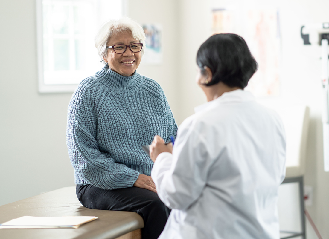 A senior woman sits up on the exam table at a doctors appointment. She is dressed casually in a blue sweater and has a smile on her face as she glances at the doctor. Her female doctor is wearing a white lab coat and seated in front of her as she takes notes on a tablet.