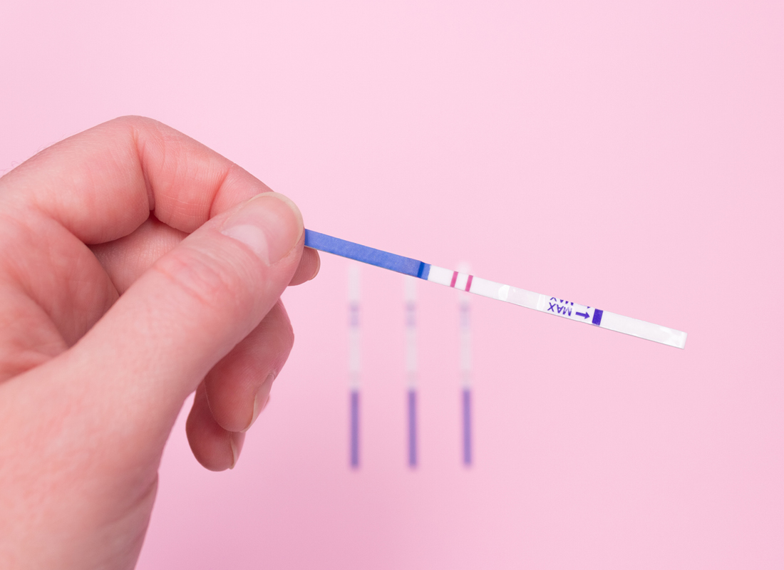 A hand holds a positive pregnancy test on a pink background