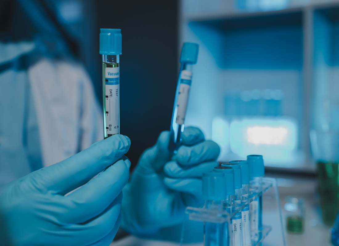A closeup of a laboratory professional in a darkened laboratory holding up two test tubes at a bench with a rack of more test tubes, illustrating a COVID-19 testing concept.
