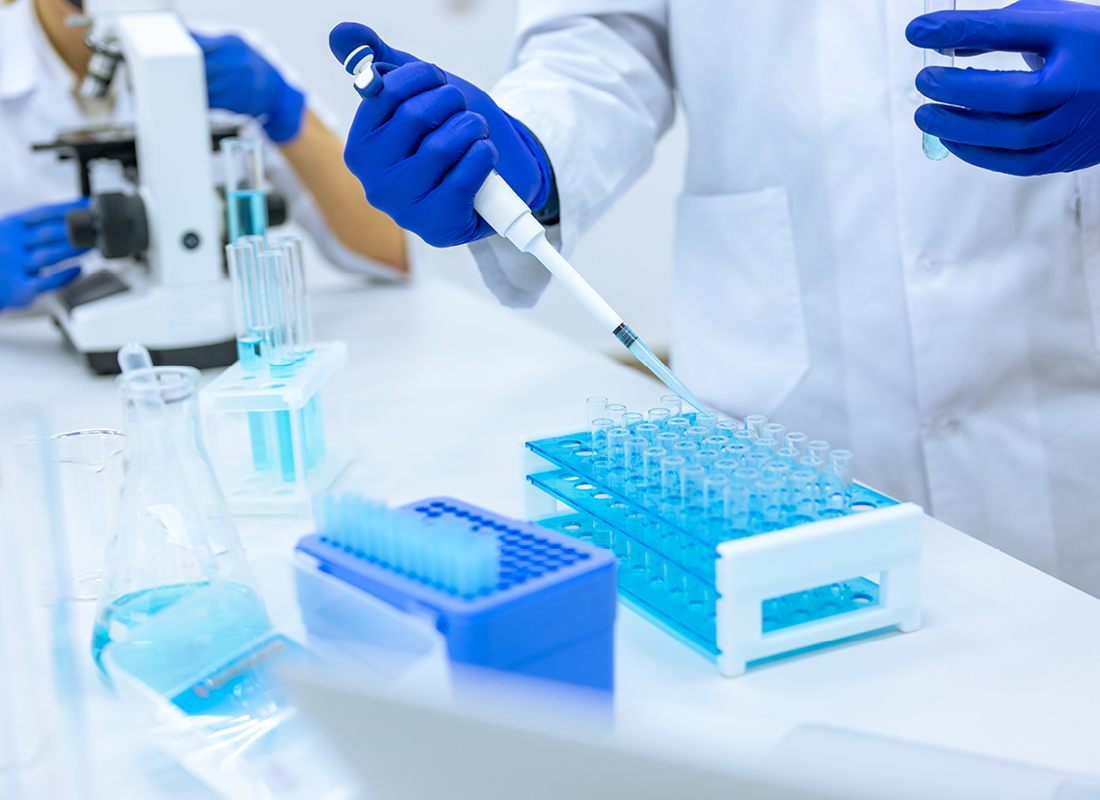 Closeup of unrecognizable scientist in surgical blue gloves dripping blue liquid from pipette into test tube at bench in laboratory.