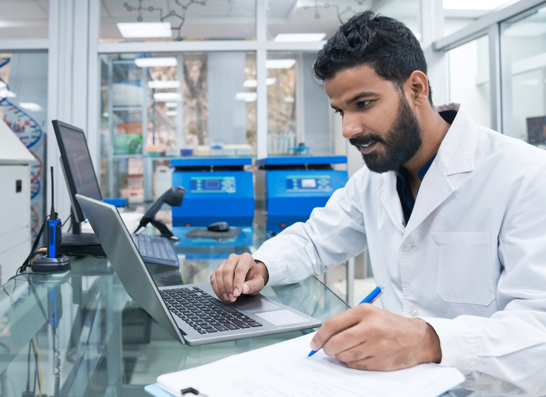 A young male laboratory professional in a lab coat uses laptop while writing on clipboard at the laboratory bench.